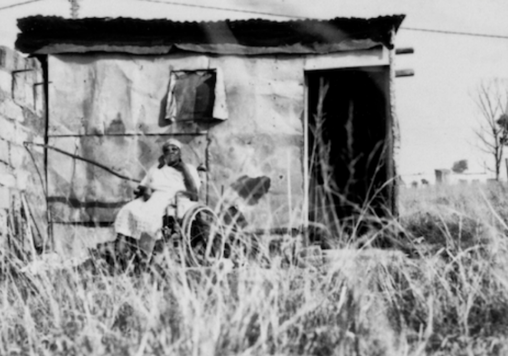 Photo of a woman in a wheelchair outside a disintegrating house.