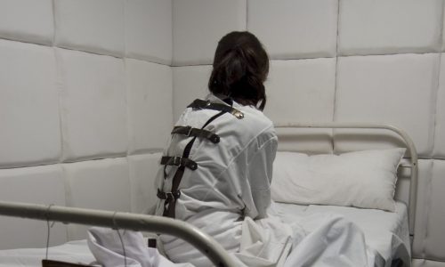 Greyscale photo of a woman sitting on a bed in a dark room.