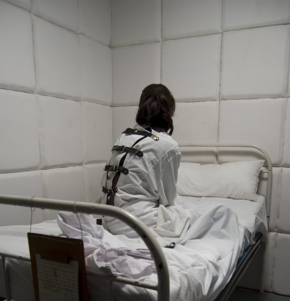 Greyscale photo of a woman sitting on a bed in a dark room.