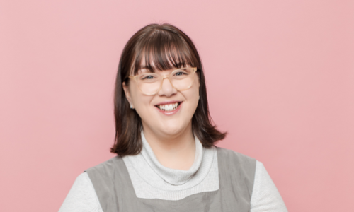 Portrait style photo of WWDA staff member, Mali Hermans. A young woman with shoulder length brown hair wearing a white top, with a grey tunic and glasses.