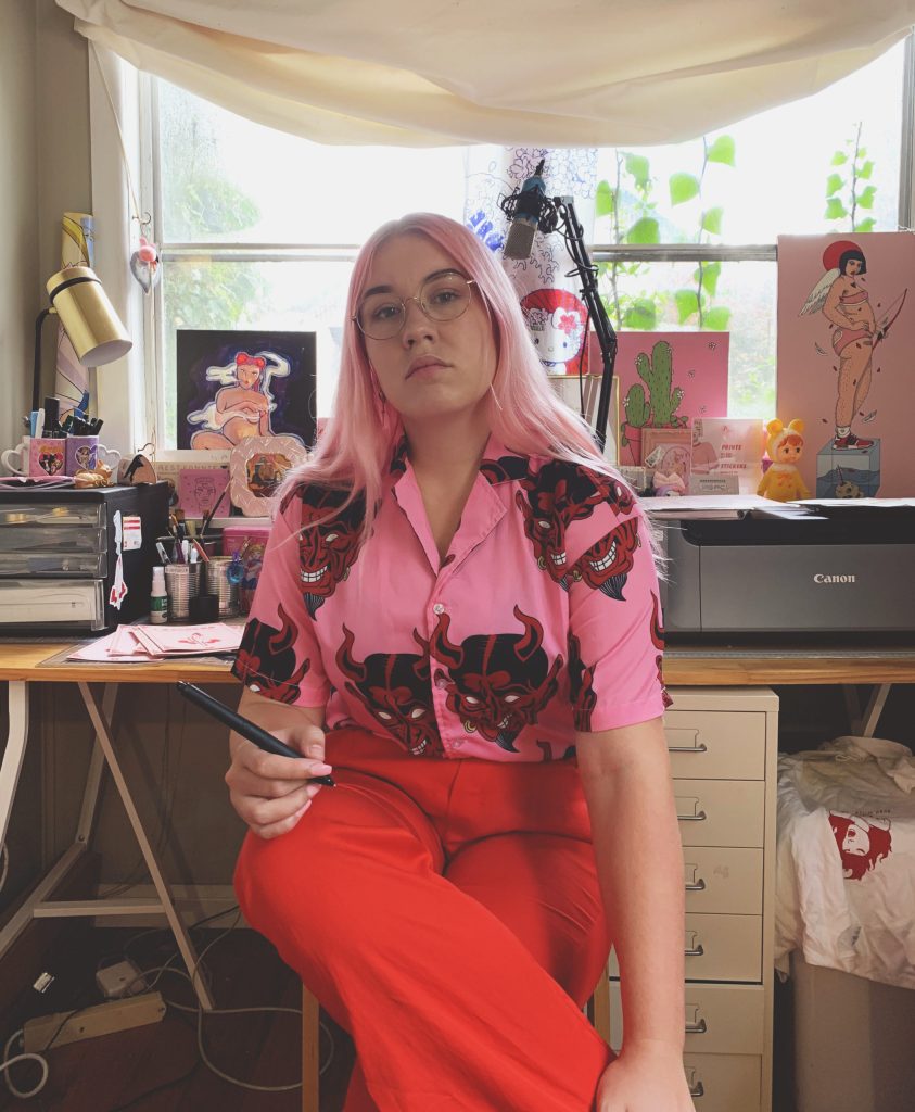 Photo of Ailie Banks, a young woman with pink hair, glasses, wearing a bright pink patterned top and bright red trousers sitting at her computer desk.