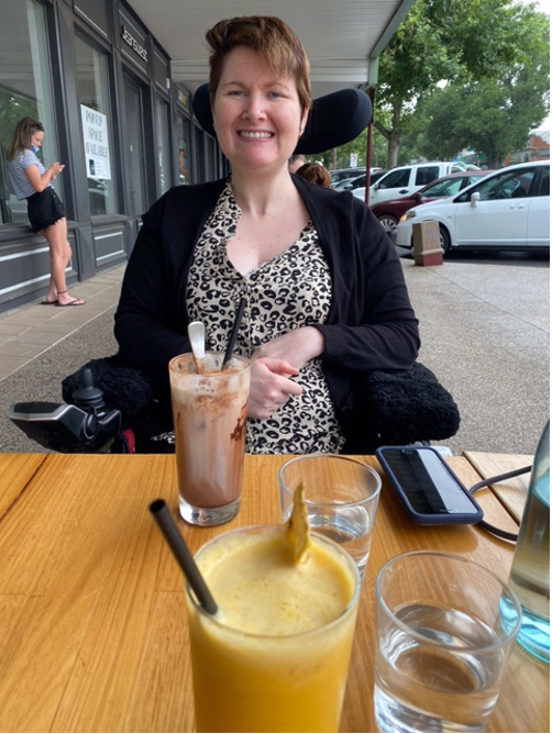 A more recent image of Anj Barker OAM sitting in her wheelchair at a cafe smiling. 