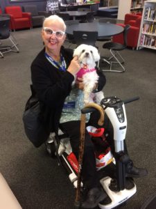 Photo of Tammy Milne cuddling a white dog wearing white glasses and red lipstick sitting in an electric wheelchair. 