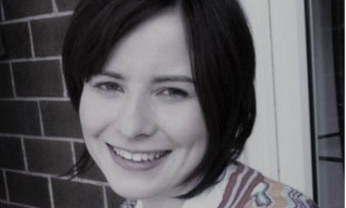 A photo of Natalie with short dark brown hair, smiling in front of a brick wall.