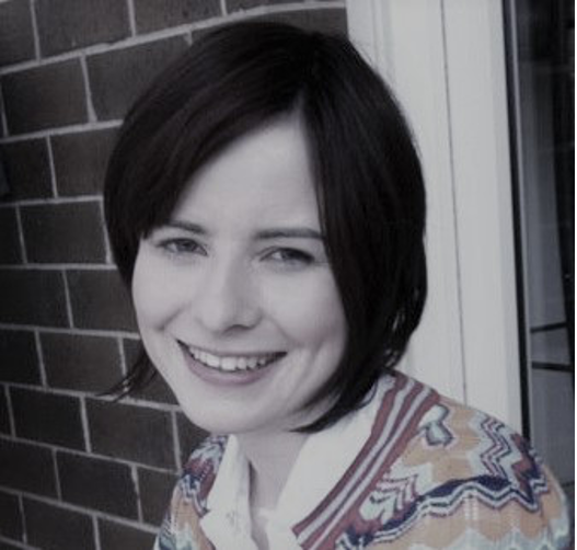 A photo of Natalie with short dark brown hair, smiling in front of a brick wall.