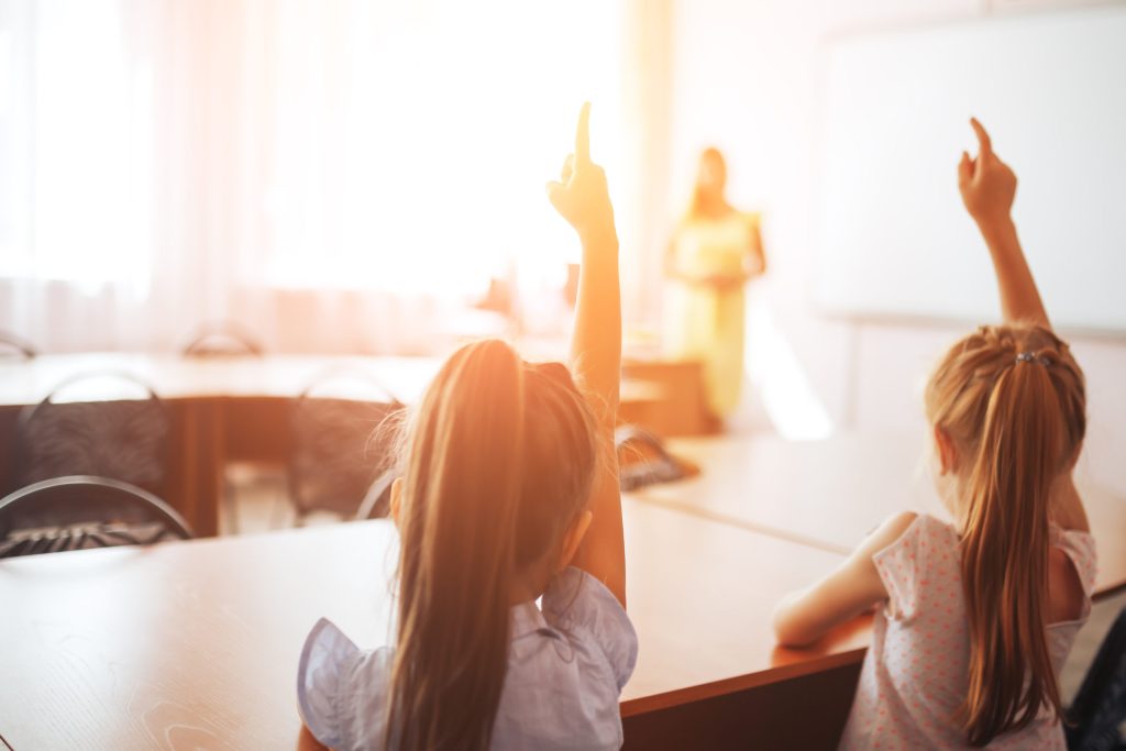 A photo of two young girls with their hands raised in a classroom