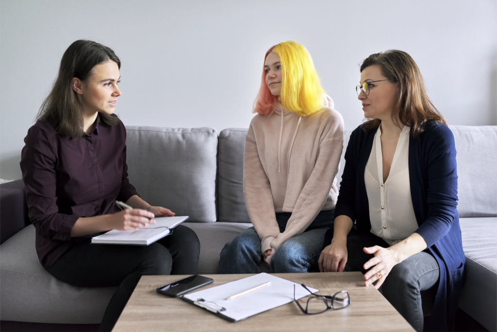 A photo of three woman sitting on a couch. One has a paper and pen facing the other two woman. One of the woman is younger with brightly coloured hair.