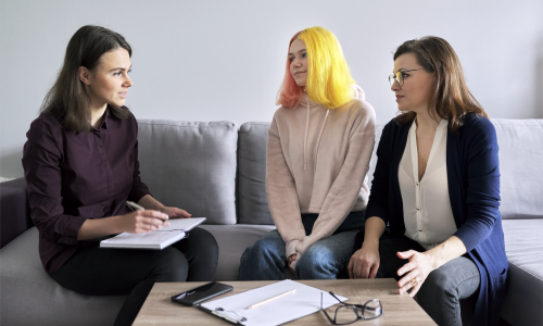 A photo of three woman sitting on a couch. One has a paper and pen facing the other two woman. One of the woman is younger with brightly coloured hair.