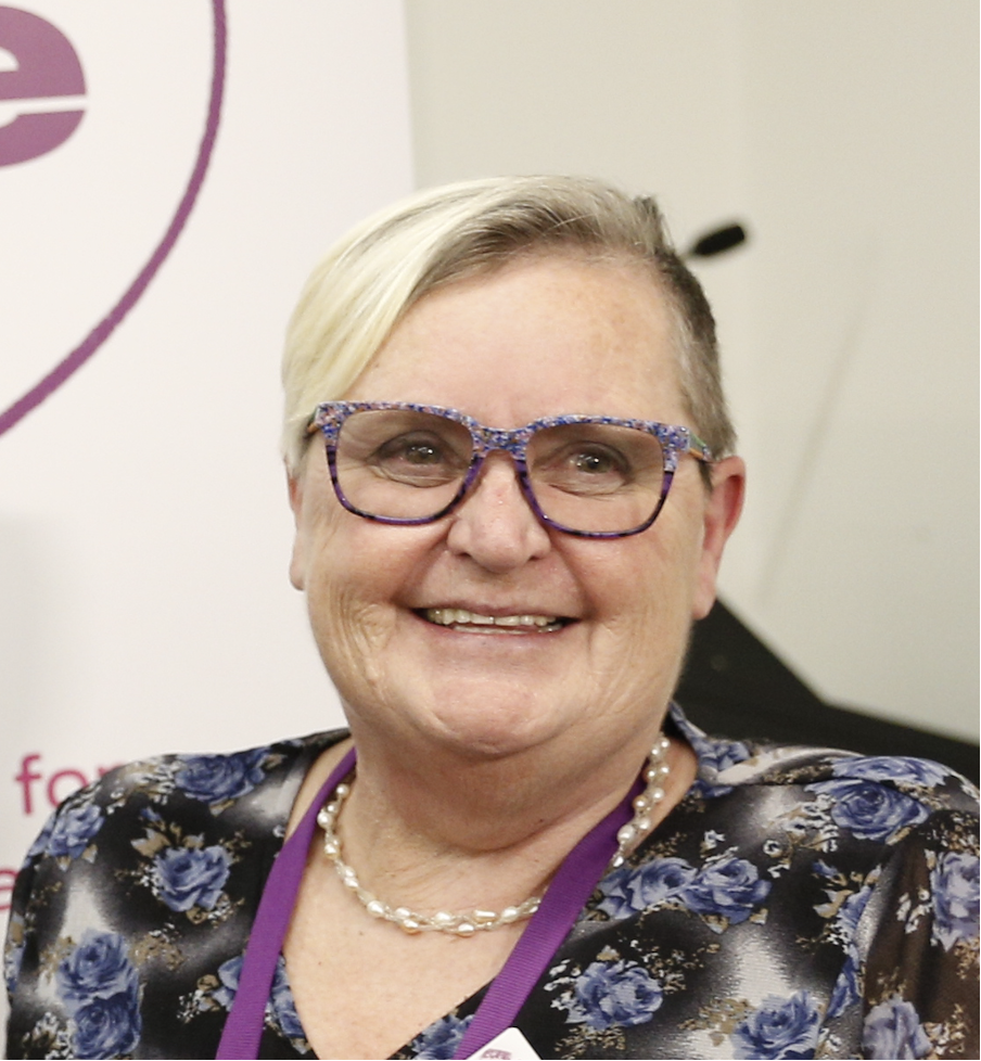 Photo of Tricia Malowney, a woman with short blonde hair and wearing purple glasses.