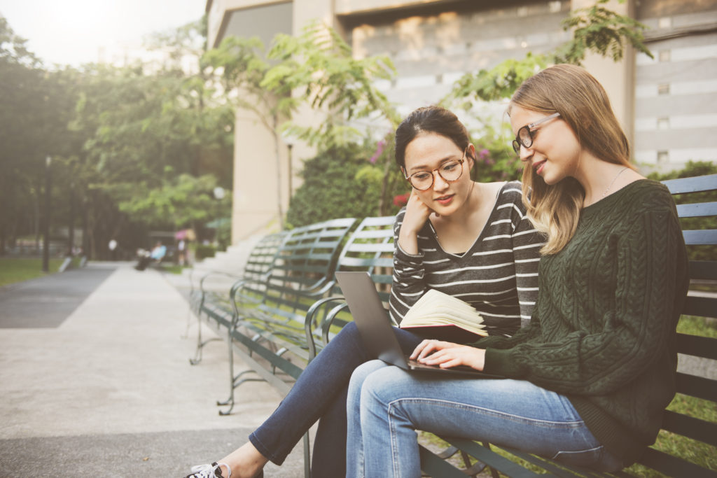 Two women who are friends sitting on a park bench, one is using a laptop and the other is reading. 