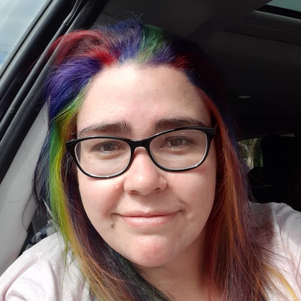 Me, embracing my difference, now.
A selfie of an Indigenous woman wearing a pink tshirt, and black rimmed glasses. She has rainbow coloured shoulder length hair.