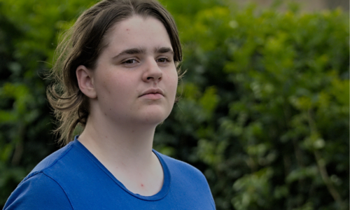 Photo of Maria who has dark brown hair tied back in a ponytail and is wearing a blue shirt that reads I love someone who is autistic (it's me).