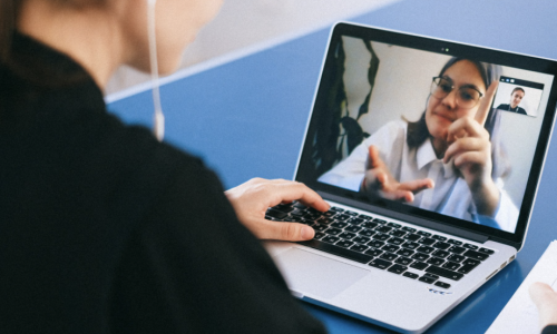 A young woman using a laptop with headphones in. On the screen is another young woman talking in Auslan.