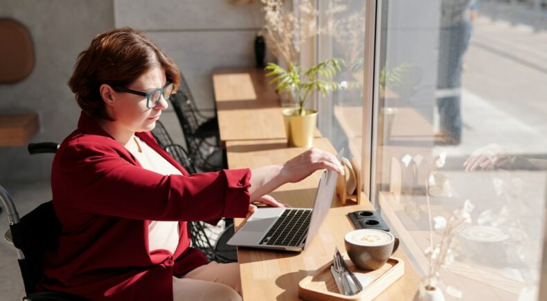 A woman wearing a white shirt and red jacket sitting at a bench in a window looking at her laptop