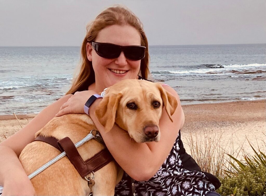 Photo of Katie who has long blonde hair and is wearing dark sunglasses. She is hugging her dog called sadie who is wearing a mobility support harness.