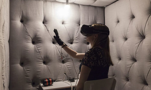 Image description: In a grey sound-proofed room with dim lighting, a woman sits at a desk. She is wearing a virtual reality headset and holding up her hand, which is wearing a custom-built glove. On the desk are different cables and speakers.