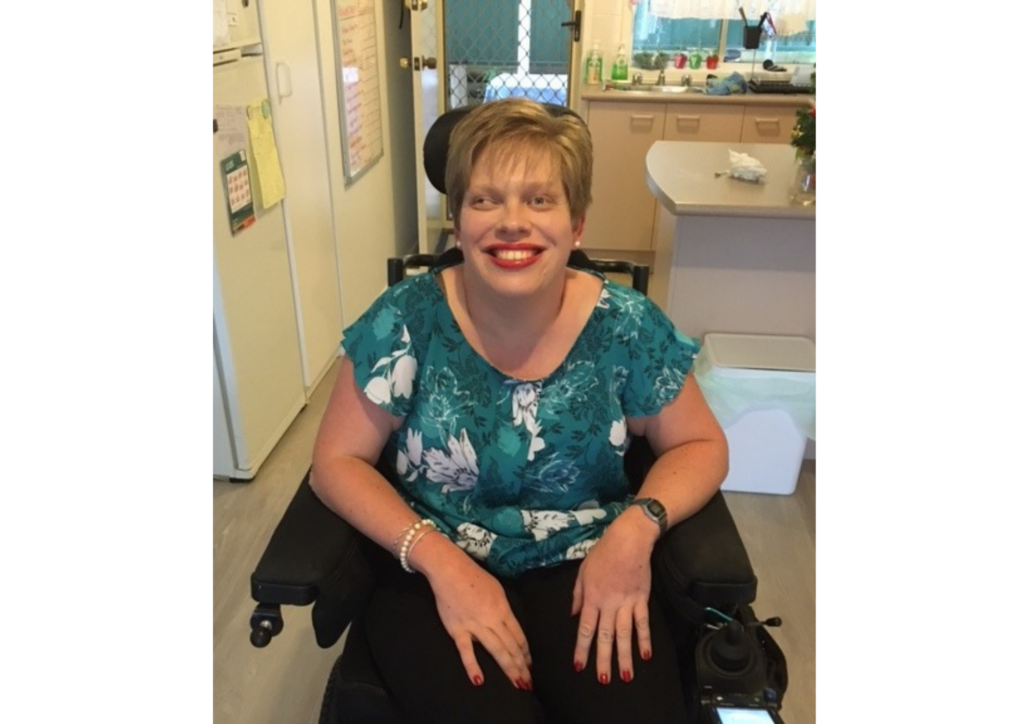 A photo of Melanie Schlaeger with short hair, red lipstick and a flowery blue top sitting in a wheelchair.