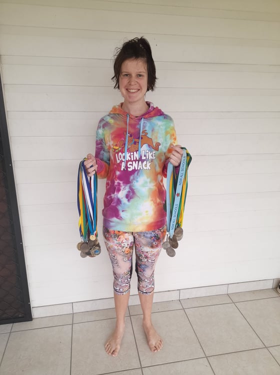 • A photo of Steph, a white woman with short brown hair, holding dozens of sporting medals. Steph is wearing a tie-dye hoodie and colourful leggings.