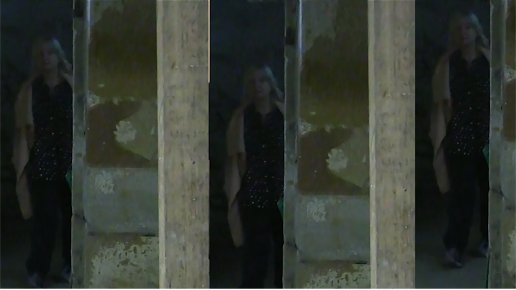 Beyond a blurred wall – which could be interpreted as bars or mirrors – is the figure of Sandie. She is obscured, but you can make out her shoulder-length grey hair.