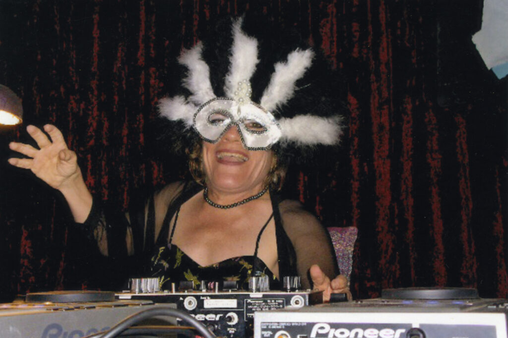 A small-statured woman wearing a black dress, see-through cardigan and white dress-up mask with feathers is smiling and laughing. She is sitting behind some DJ decks she is using at a masked-ball event.