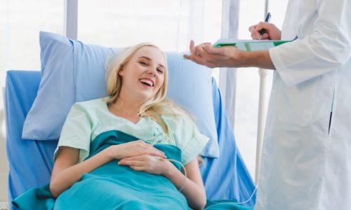 A doctor visiting a young woman in a hospital bed. The doctor is holding a clipboard and talking to the young woman.