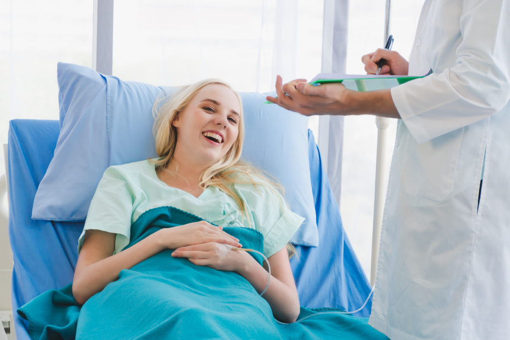 A doctor visiting a young woman in a hospital bed. The doctor is holding a clipboard and talking to the young woman.