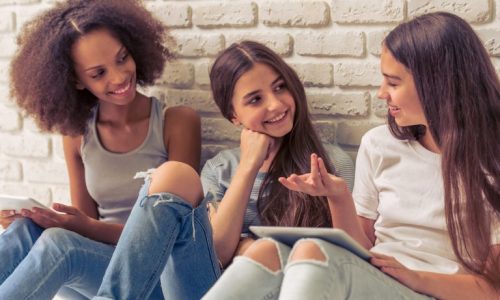 Three young women sitting against a wall talking.