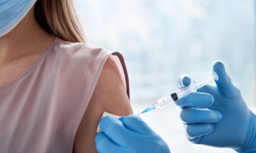 An image of a woman being injected with a COVID vaccine by a doctor wearing blue sterile gloves. Only the woman’s shoulder and neck are visible. She has white skin, blonde hair and is wearing a light pink, sleeveless blouse.