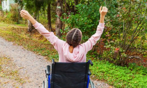 Photo of a young woman using a wheelchair. She is wearing pink and has her arms up in the air.