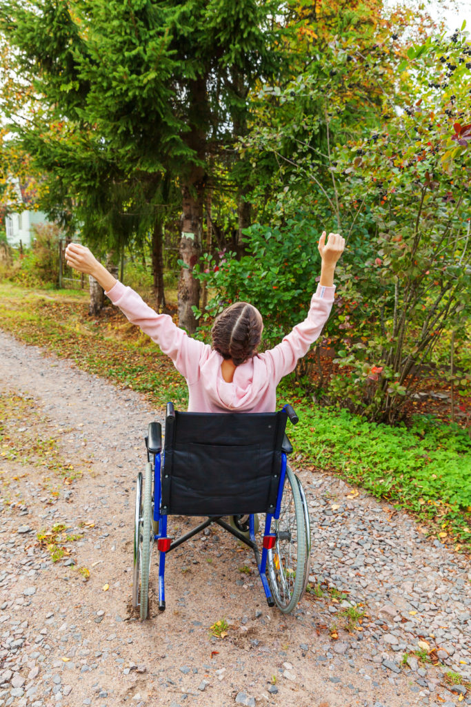 Photo of a young woman using a wheelchair. She is wearing pink and has her arms up in the air.