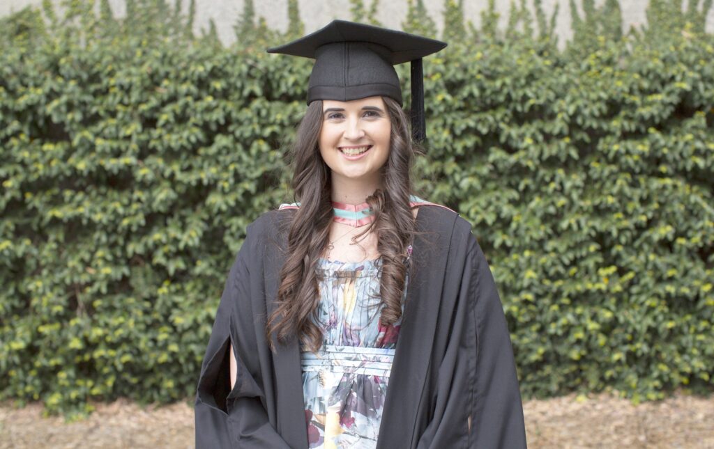 An image of Katherine smiling in front of greenery in graduation attire. Katherine has long brown hair that is curled, and is wearing a colourful floral dress, black graduation robe and black graduation cap.