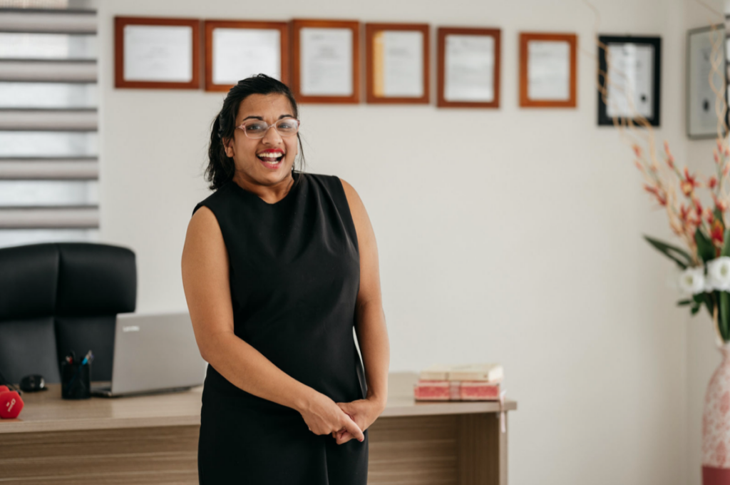 A photo of Jerusha standing and smiling at the camera. Jerusha has brown skin and black hair in a ponytail. She is wearing a black dress and glasses. In the background you can see an office desk, chair and laptop, as well as qualifications and degrees hanging on the wall.