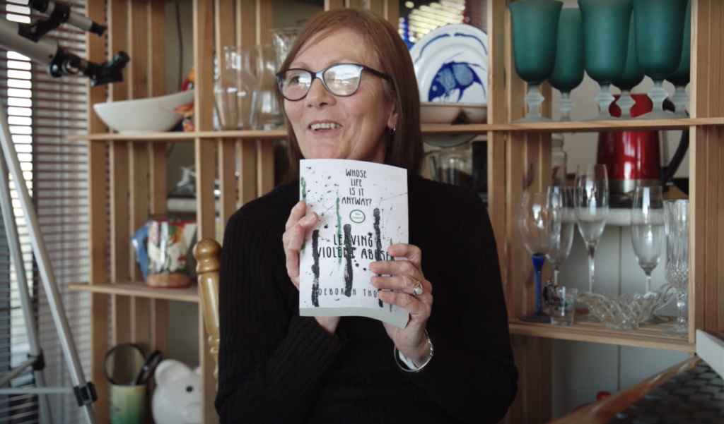 A photo of Deb Thomson holding a copy of her book 'Whose life is it anyway? Leaving a violent abuser' and smiling. Deb is a white woman with black glasses and shoulder length brown hair. She is wearing a black turtle-neck shirt. In the background is a cabinet with glasses, fine china and a telescope.