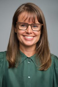 A headshot of Bonney, smiling at the camera against a grey backdrop. She has fair skin, shoulder length light brown hair and blue eyes. Bonney is wearing brown, square-framed glasses and an emerald green collared-blouse.  