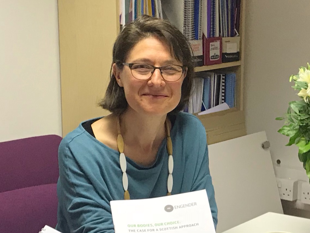 A photo of Jen smiling with a closed mouth at the camera. She has shoulder length dark brown hair and fair skin, is wearing dark-framed glasses, a blue long-sleeved shirt and a necklace with elongated green and white wooden beads. She is sitting on a purple office chair with a cabinet of files behind her, a vase of flowers to the right and a white sign in front of her that reads “Engender. Our Bodies, Our Choice. The case for a Scottish Approach.”