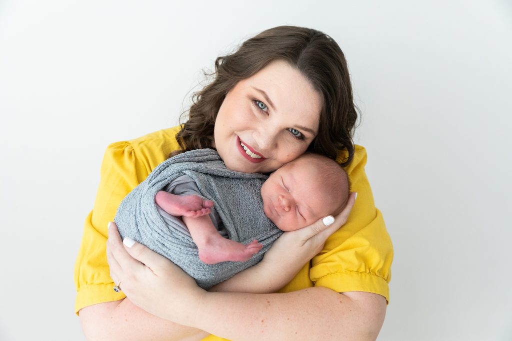 A photo of Annie smiling at the camera and cradling her newborn baby. Annie is a white woman with shoulder length brown hair wearing a bright yellow shirt. Her baby is wrapped in a blue-grey swaddle.