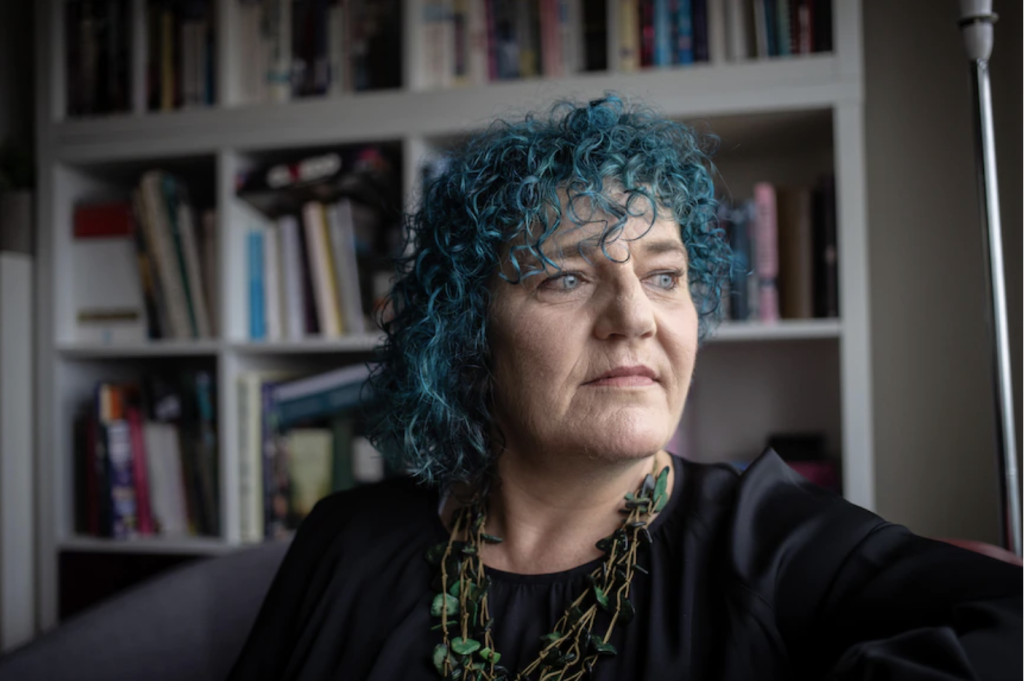 Photo of WWDA CEO Carolyn Frohmader. Carolyn has blue, short, curly hair and is looking out a window wearing a black top and green necklace in front of a book shelf.