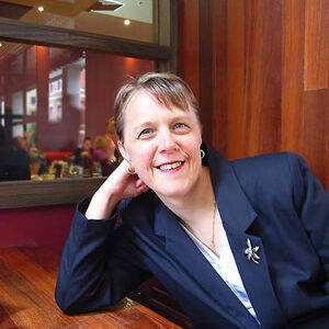 A photo of Kerryn sitting at a wooden table, leaning their head on one hand and smiling at the camera. Kerryn has white skin, short mousy brown hair, and is wearing a navy blazer and brooch over a light blue blouse.