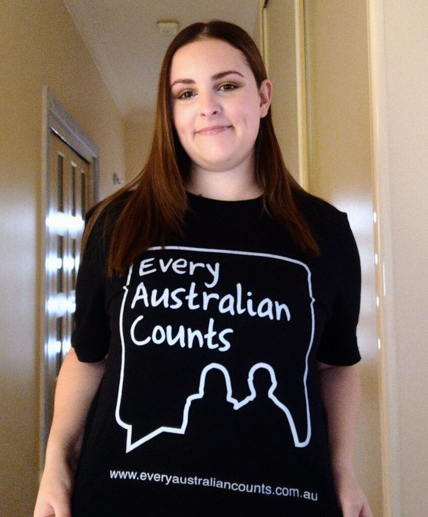 A photo of Bethany, smiling with her mouth closed and holding her black and white Every Australian Counts #DefendOurNDIS campaign shirt so it is visible. She has fair skin, long brown hair, and is standing in the hallway of a house.