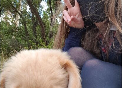 An image of a young Autistic person with fair skin and long light brown hair. They are wearing a black face mask and doing a peace sign with one hand. They are sitting down with their knees to their chest, wearing navy stockings. Leaning on their legs is Goose, a golden retriever service dog. In the background are trees.