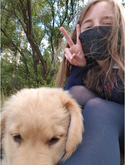 An image of a young Autistic person with fair skin and long light brown hair. They are wearing a black face mask and doing a peace sign with one hand. They are sitting down with their knees to their chest, wearing navy stockings. Leaning on their legs is Goose, a golden retriever service dog. In the background are trees.