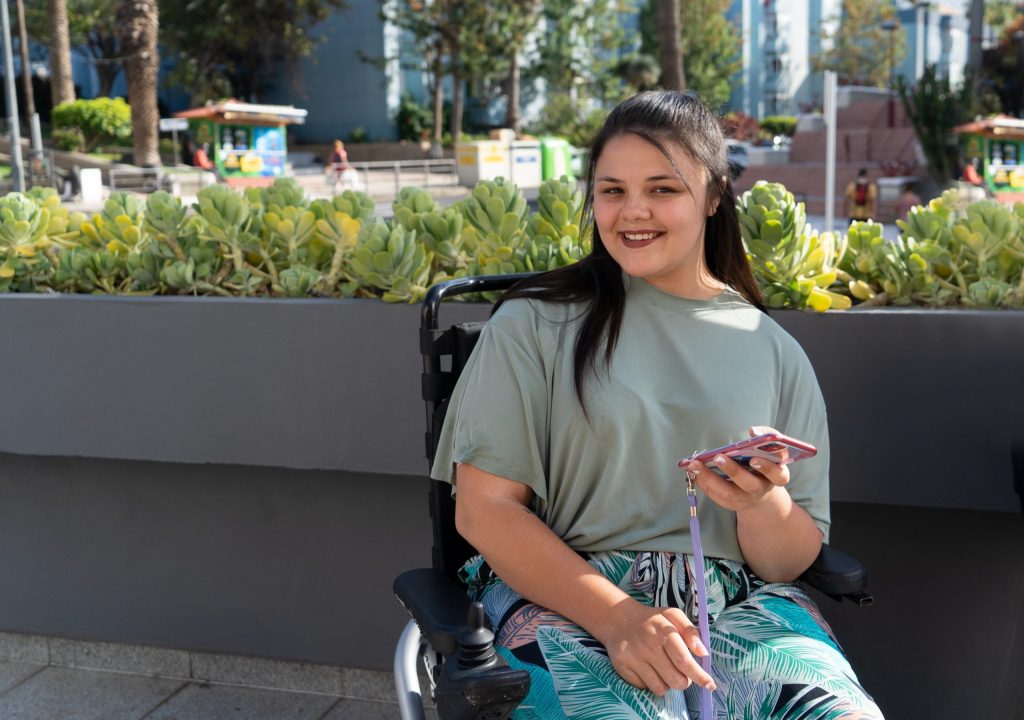 Young woman using a wheelchair smiling at the camera and holding up a smart phone. In the background is a concrete planter with green plants in it.