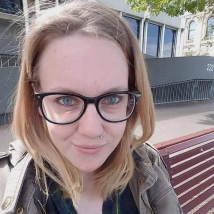 A selfie of Ferris smiling with their mouth closed at the camera. They have white skin, shoulder length blonde hair, blue eyes, and a silver nose ring. They are wearing a green khaki jacket, a black shirt, black-rimmed glasses and are sitting on a bench in front of a city building.