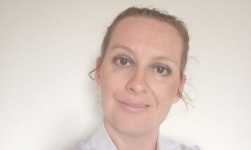 A portrait photo of Kerry who has white skin and light brown hair tied back and she is smiling. Kerry is wearing a white button up shirt.