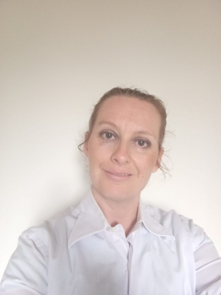 A portrait photo of Kerry who has white skin and light brown hair tied back and she is smiling. Kerry is wearing a white button up shirt.