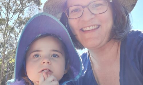 Photo of Marie and Izzy. Marie-Christine outside with daughter Izzy. They have white skin and both are wearing patterned tops and hats. Marie-Christine is wearing glasses and smiling. Izzy has her hands clasped together raised near face.