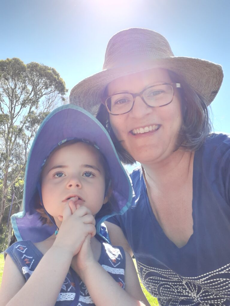 Photo of Marie and Izzy. Marie-Christine outside with daughter Izzy. They have white skin and both are wearing patterned tops and hats. Marie-Christine is wearing glasses and smiling. Izzy has her hands clasped together raised near face.