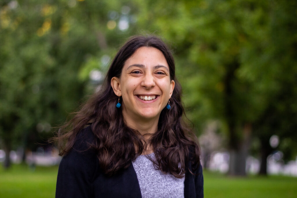 A head shot of Laura outdoors, with a massive smile on her face. Laura has white skin and long wavy dark brown hair. She is wearing a grey top, a black cardigan and round turquoise drop earrings.