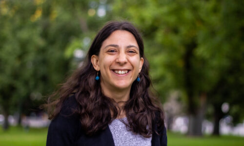 A head shot of Laura outdoors, with a massive smile on her face. Laura has white skin and long wavy dark brown hair. She is wearing a grey top, a black cardigan and round turquoise drop earrings.