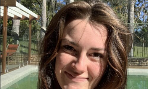 Portrait photo of Breagh smiling, outside with a pool in the background. She has white skin and very long brown hair and is wearing a white tank top.